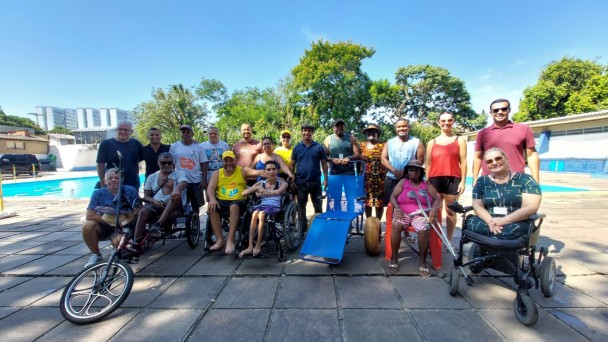 Foto: ao fundo da Piscina do Cevi, a frente em primeiro planom o presidente da Faders ao lado da cadeira anfíbia e demais cadeirantes que estavam no evento, em pés atrás deles as demais autoridades e famílias que participaram da entrega da cadeira que agora será utlizada para banhos assistidos para pessoas com deficiência e pessoas com mobilidade reduzida. Fim da descrição