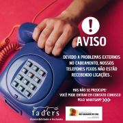 Card com fundo vermelho, no canto esquerdo ha um telefone azul fixo com uma mão segurando o gancho- do lado direito no topo a imagem de aviso e escrito em abaixo "AVISO"-  segue o texto: Devido a problemas externos no cabeamento, nossos telefones fixos não estão recebendo ligações-  Mas não se preocupe! Você pode entrar em contato conosco pelo Whatsapp. Abaixo ainda destes textosas as logos da Faders e da Sec. Desenvolvimento Social RS . Fim da descrição