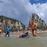 Foto- dois guarda vidas acompanham uma senhora na cadeira anfíbia em um banho de mar.
