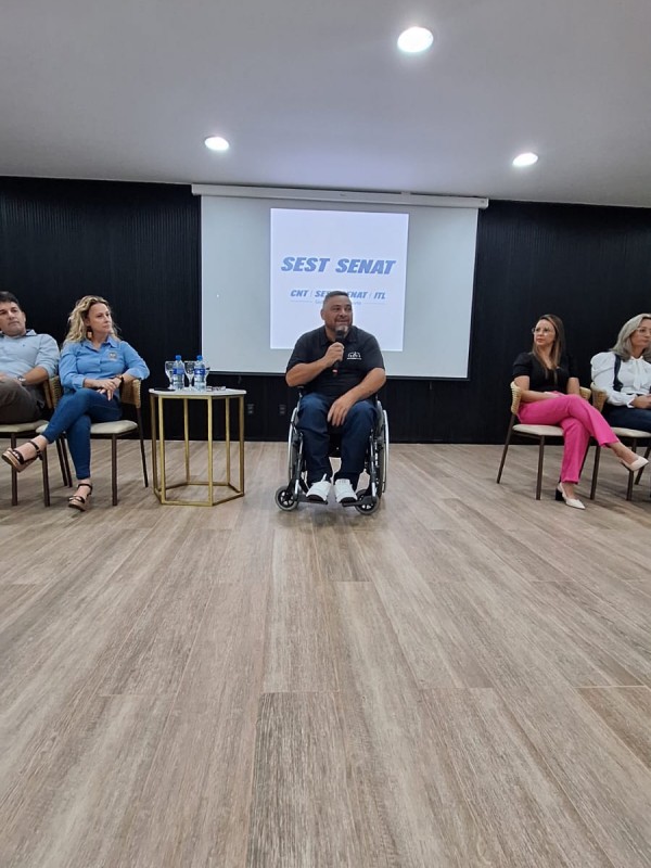 Foto- ao centro o presidente da Faders em sua cadeira de rodas com o microfone em mãos faz sua fala na abertura do evento, ao fundo um telão com as logos do Sesc-senat ao lado esquerdo uma mesa bege com garrafas e copos de água em cima ao lado em cadeiras beges a prefeita, o promotor....do lado direito também cadeiras beges e as demais autoridades presentes no evento. Fim da Descrição 