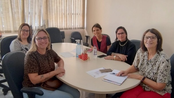 Foto: paredes em tom de um pessego placo, mesa com tampa bege clara madeira e cadeiras com encostos pretos ao redor, nelas a vereadora de Bagé, Ana Paula da Silva Moreira, a assessora jurídica Marília Scardoelli dos Santos, a assessora parlamentar Maíra Zago e a intérprete de Libras Renata de Melo Machado, todas de Bagé, e a coordenadora de acessibilidade da Faders, Jaqueline Rosa 