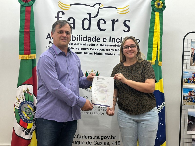 foto, ao fundo o banner da Faders, com as bandeiras do RS e Brasil, em frente o chefe de gabinete da Faders recebendo a carta intenções Bagé da Vereadora. Fim da descrição