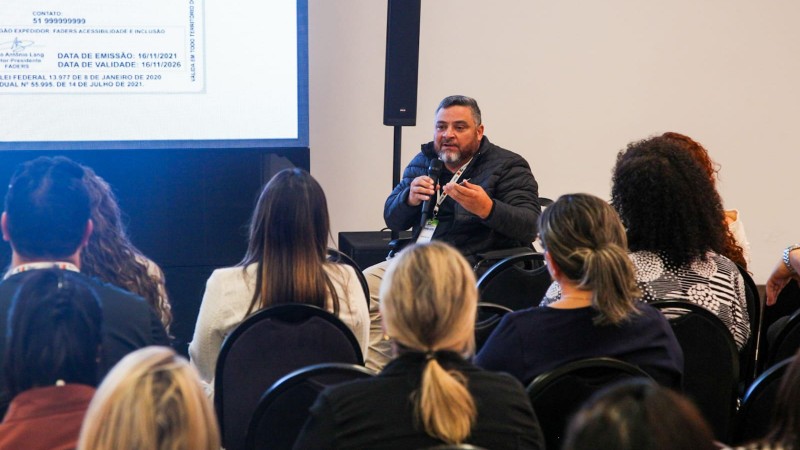 foto - publico de costas sentado em cadeiras com encostos preto, e em frente a uma tela onde está a apresentação da Ciptea, o presidente da Faders, em sua cadeira de rodas, e com o microfone em uma das mãos e a Carteira de identificação da Pessoa com Especto Autista em outra, fala sobre a importância do documento. Fim da  descrição