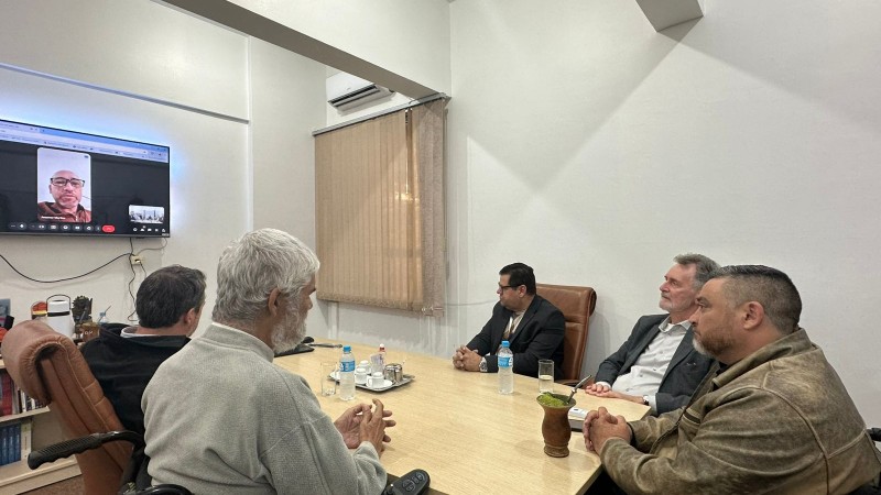 Foto- sala com paredes brancas, ao centro uma mesa com taqmpa em madeira clara, ao redor cadeiras em couro marrom, na ponta da mesa em sua cadeira de rodas o presidente da Faders, a dua esquerda o presidente do Coepede também em sua cadeira de Rodas, ao redor deles os demais três participantes da reunião. Em frente a mesa uma televisao transmite a participação por videoconferência de um delegado do Coepede. Fim da descrição