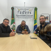 foto- ao fundo parede branca, com o banner da Faders e as bandeiras do RS e Brasil, mais a direita da pra ver um painel de fotografias com a sprincipais atividades da Fundação. Em primeiro plano ao redor de uma mesa com tampa de madeira clara esta na ponta o presidente da Faders em sua cadeira de Rodas, do lado esquerod o diretor geral em uma cadeira com encosto marrom em couro e do direito o diretor de inclusão também em sua cadeira de rosas  da Secretaria de Justiça, Cidadania e Direitos Humanos do Rio Grande do Sul. Fim da Descrição