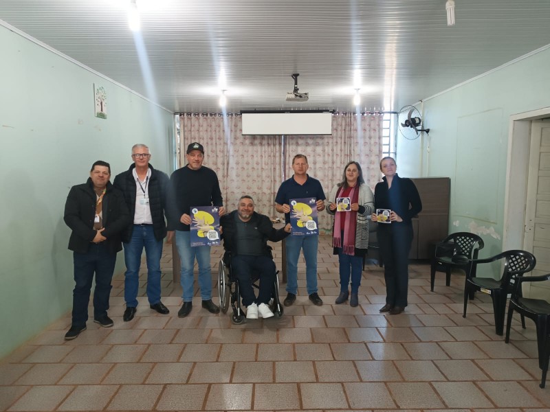 Foto - em uma sala com paredes verdes e piso bege claro, em pé estão 6 pessoas e ao centro o presidente da Faders em sua cadeira de rodas. Fim da descrição