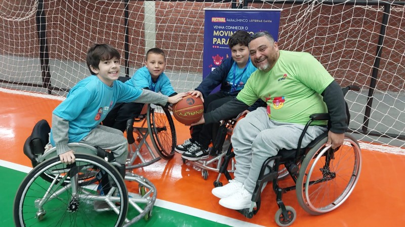 FOTO- em uma quadra de esportes estão o presidente da Faders e mais 3 meninos com idades variadas em suas cadeiras de rodas em um circulo e ao centro eles seguram uma bola de basquete. Os meninos estão de camiseta azul e abrigo cinza e o presidente da Faders de camiseta verde e abrigo cinza. Todos sorriem com expressão de felicidade. Fim da descrição