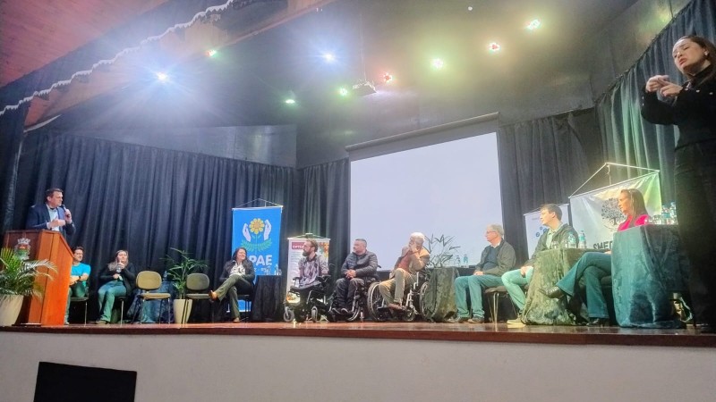 Foto - Do placo do evento onde estão sentadas em cadeiras pretas as autoridades que participaram da abertura oficial do evento. Entre elas em suas cadeiras de rodas o presidente da Faders e o presidente do Coepede. Na tribuna o prefeito de Feliz faz sua fala com op microfone em maos e vestindo uma camisa e blazer azul. Fim da descrição