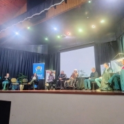 Foto - Do placo do evento onde estão sentadas em cadeiras pretas as autoridades que participaram da abertura oficial do evento. Entre elas em suas cadeiras de rodas o presidente da Faders e o presidente do Coepede. Na tribuna o prefeito de Feliz faz  sua fala com op microfone em maos e vestindo uma camisa e blazer azul. Fim da descrição 