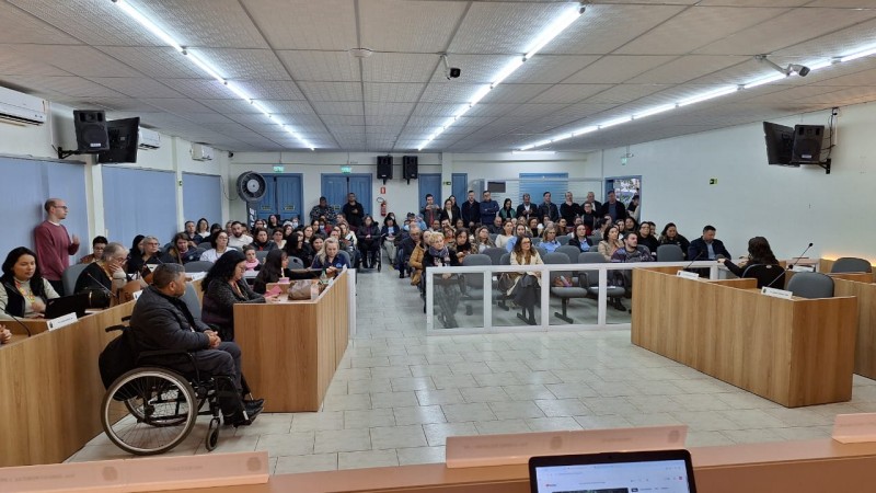 Foto- auditório da câmara de Vereadores de Santo Antônio da Patrulha, foto tirada da mesa da presidencia, que pega de frente as mesas dos vereadores onde está a equipe da Fader e o presidente, em frente as cadeiras  azuis dos visitantes onde estão os participantes do evento sentados, e alguns em pé no fundo. A sala está lotada. Fim da descrição