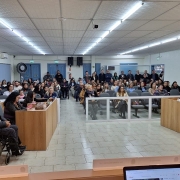 Foto- auditório da câmara de Vereadores de Santo Antônio da Patrulha, foto tirada da mesa da presidencia, que pega de frente as mesas dos vereadores onde está a equipe da Fader e o presidente, em frente as cadeiras  azuis dos visitantes onde estão os participantes do evento sentados, e alguns em pé no fundo. A sala está lotada. Fim da descrição