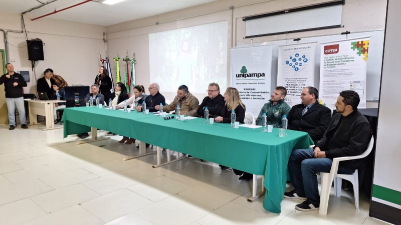 Foto- Paredes brancas e piso claro, ao fundo um telão, e os banners da Faders e da Unipampa. Em primeiro plano a mesa das autoridades com uma toalha verde bandeira, as cadeiras são brancas. Mesa, estão as autoridades que participaram da mesa de abertura do 206º Fórum Permanente que aconteceu em Bagé. No centro da mesa o presidente da Faders e o prefeito de Bagé. No canto esquerdo em pé um interprete de libras. Fim da descrição