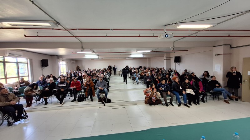 Foto- Paredes brancas e piso claro, ao fundo as cadeiras do auditório com encosto azul, no meio delas um corredor, a sala esta lotada e tem pessoa em pé ao fundo. Fim da descrição
