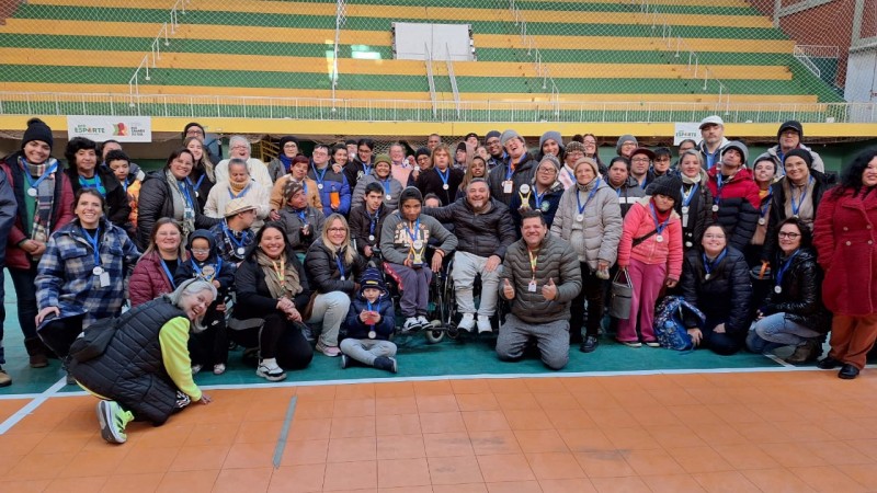 Foto- no estádio Militão, ao fundo aparecem as arquibancadas em verde e amarelo, na quadra já está o grupo que participou do Desafie-se Faders alguns em pés e uns no chão sentados ou acocorados. Todos com suas medalhas de participação, muito sorridentes, entre eles além dos alunos, professores, familiares e equipe de Bagé da faders que organizou o evento. No centro o presidente da Faders em sua cadeira de rodas e os representantes das instituições com maior número de participantes que tem em mãos o troféu recebido. Fim da descrição