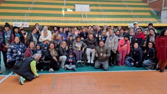 Foto- no estádio Militão, ao fundo aparecem as arquibancadas em verde e amarelo, na quadra já está o grupo que participou do Desafie-se Faders alguns em pés e uns  no chão sentados ou acocorados. Todos com suas medalhas de participação, muito sorridentes, entre eles além dos alunos, professores, familiares e equipe de Bagé da faders que organizou o  evento. No centro o presidente da Faders em sua cadeira de rodas e os representantes das instituições com maior número de participantes que tem em mãos o troféu recebido. Fim da descrição