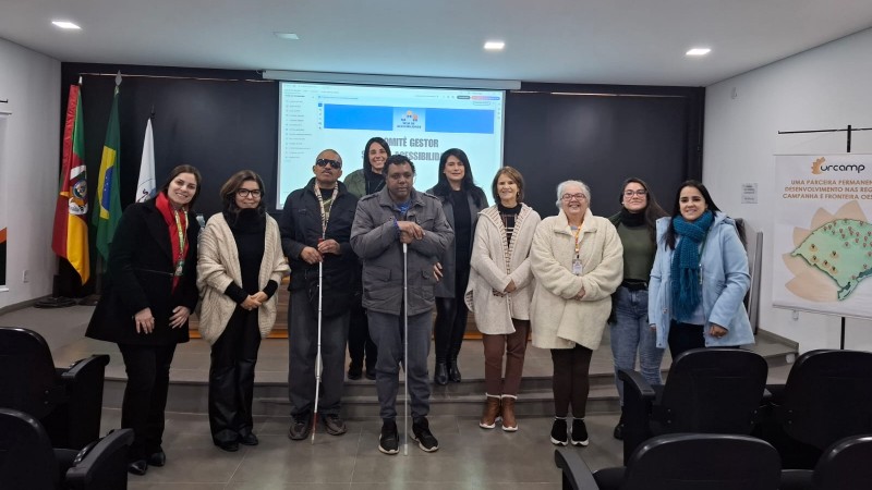 Foto - sala com paredes brancas, ao fundo parede preta com um telão no centro onde esta sendo projetado o material debatido na Oficina. Na frente em primeiro plano e todos em pé, a equipe que faz parte do Comitê Gestor Municipal do Sele de Acessibilidade em Bagé, a diretora técnica e a coordenadora de Acessibilidade, ambas da Faders. Fim da descrição