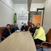 Foto - Cinco pessoas estão sentadas em torno de uma mesa retangular de madeira clara, numa sala de reuniões. As cadeiras são em couro marrom. Ao fundo, há uma faixa institucional da FADERS – Acessibilidade e Inclusão, com as bandeiras do Brasil e do Rio Grande do Sul ao lado. Todos estão sorrindo para a foto.
Da esquerda para a direita:
1.	O diretor administrativo Faders, de terno preto com crachá;
2.	A diretora Técnica da Faders, de cabelo grisalho e óculos, vestindo colete jeans;
3.	Um homem ao centro, de casaco preto, com as mãos apoiadas na mesa (presidente Marquinho Lang);
4.	Novos intérpretes: Uma mulher de cabelo castanho claro e ondulado, com casaco preto (Sharon); 5 -Um homem de barba curta, usando moletom amarelo com estampa (Andrei).
O ambiente é iluminado por lâmpadas de teto, e no canto superior direito há um ventilador de teto visível.

