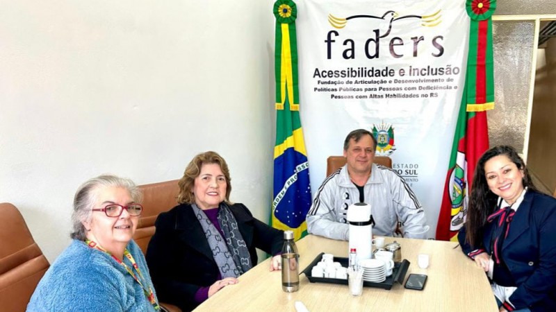 Foto - ao redor de uma mesa de tampa clara estao o chefe de gabinete da Faders, e a diretora técnica da faders, reunidos com as gestoras de NH, ao fundo o banner da Faders e as bandeiras do Brasil e do RS.