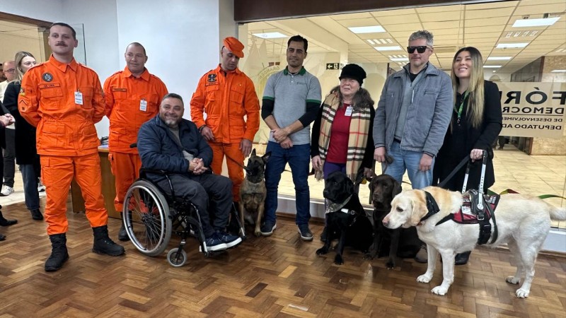 Um grupo de pessoas posa para foto em no espa&ccedil;o de Converg&ecirc;ncia da ALRS . &Agrave; esquerda, tr&ecirc;s homens vestem uniformes laranja do Corpo de Bombeirose um deles segura poela coleira uma cachorra de plagem marrom com preto, a Melt que &eacute; c&atilde;o de resgate da equipe. Ao lado deles, o presidente da Faders em cadeira de rodas sorri para a c&acirc;mera. No centro, outro homem de p&eacute; ( professor Leonardo) e uma mulher de boina preta e cachecol xadrez est&atilde;o acompanhados por seus c&atilde;es-guia, um de pelagem preta e marrom ( Kobi e Kaiser). &Agrave; direita, um homem com &oacute;culos escuros segura um c&atilde;o-guia de marrom o Kaiser, enquanto uma mulher de cabelo comprido sorri ao lado com seu c&atilde;o-guia de pelagem clara. Todos est&atilde;o reunidos em ambiente com piso de madeira e uma parede de vidro ao fundo.Fim da descri&ccedil;&atilde;o