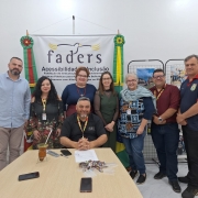 Foto posada de um grupo de sete pessoas em pé ( representantes de Vacaria, diretores da Faders da área Administrativa e Técnia, Coordenadora de Direitos e chefe de Gabinete), sorrindo, em frente ao painel da FADERS – Acessibilidade e Inclusão. À frente, o presidente da Faders está sentado à mesa com chimarrão e papéis, também sorrindo. Ao fundo, bandeiras do Rio Grande do Sul e do Brasil, além de painéis com fotos coloridas que retratam atividades e eventos.