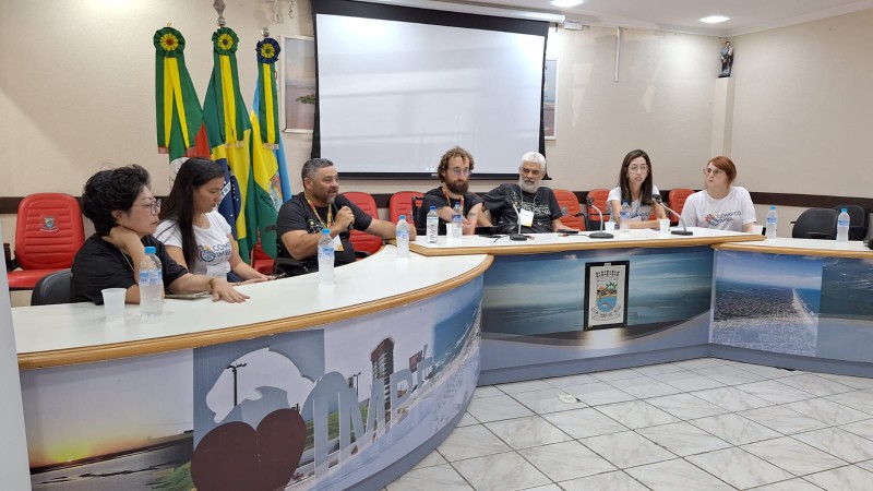 Fotografia - plen&aacute;rio da c&acirc;mara de vereadores de Imb&eacute; durante um encontro institucional sobre pol&iacute;ticas para pessoas com defici&ecirc;ncia. Na parte frontal da sala, uma mesa longa em formato semicircular re&uacute;ne cerca de oito participantes sentados, que parecem compor uma mesa de debate ou painel. Atr&aacute;s deles h&aacute; tr&ecirc;s bandeiras e um tel&atilde;o de proje&ccedil;&atilde;o ao centro da parede.

&Agrave; direita do palco, uma mulher em p&eacute; realiza interpreta&ccedil;&atilde;o em L&iacute;ngua Brasileira de Sinais (Libras). Pr&oacute;ximo a ela h&aacute; um banner grande com a sigla COMPCD IMB&Eacute;, indicando o Conselho Municipal da Pessoa com Defici&ecirc;ncia do munic&iacute;pio. &Agrave; esquerda, outro banner institucional est&aacute; posicionado ao lado de um p&uacute;lpito.

No audit&oacute;rio, v&aacute;rias pessoas est&atilde;o sentadas em cadeiras vermelhas assistindo &agrave; atividade. Entre os participantes da plateia h&aacute; pessoas com defici&ecirc;ncia, incluindo um homem em cadeira de rodas. O espa&ccedil;o possui ilumina&ccedil;&atilde;o clara, piso branco e equipamentos de ar-condicionado nas paredes, caracterizando um ambiente de reuni&atilde;o p&uacute;blica ou semin&aacute;rio.

A cena mostra um momento de discuss&atilde;o e participa&ccedil;&atilde;o social sobre direitos e pol&iacute;ticas voltadas &agrave;s pessoas com defici&ecirc;ncia. E o presidente da Faders est&aacute; com microfone em m&atilde;os realizando sua fala.
