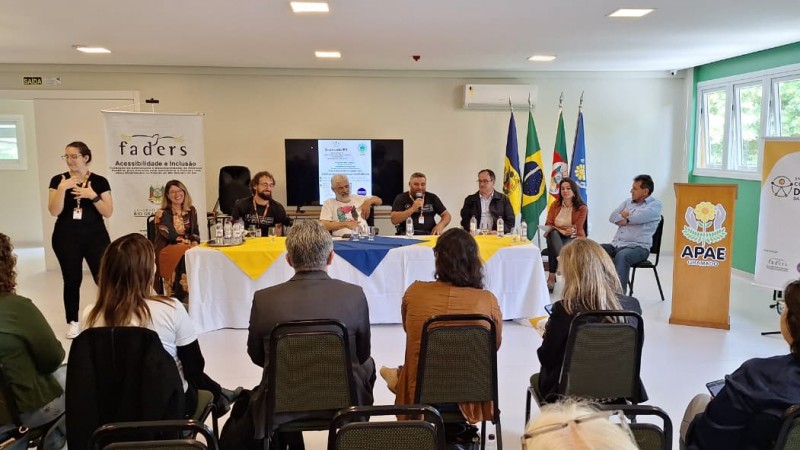 Mesa de debate com 6 participantes sentados em frente ao p&uacute;blico durante um evento institucional. O Presidente da Faders fala ao microfone enquanto os demais acompanham a discuss&atilde;o. A mesa est&aacute; coberta com toalhas nas cores branco, azul e amarelo, com copos e garrafas de &aacute;gua. Ao fundo aparecem bandeiras oficiais e um tel&atilde;o exibindo a programa&ccedil;&atilde;o do encontro. &Agrave; esquerda est&aacute; um banner da Faders Acessibilidade e Inclus&atilde;o e a interprete de libras. O publico estra em cadeirtas pretas acompanhando a fala.