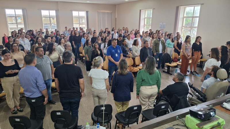 Fotografia em ambiente interno, no audit&oacute;rio da C&acirc;mara de Vereadores de Santiago, com grande n&uacute;mero de pessoas reunidas. A imagem mostra dezenas de participantes em p&eacute;, organizados entre fileiras de cadeiras, voltados para a parte frontal da sala.
No primeiro plano, aparecem algumas pessoas de costas, possivelmente autoridades ou organizadores, posicionadas &agrave; frente do p&uacute;blico. Entre elas, h&aacute; um homem utilizando cadeira de rodas. Tamb&eacute;m &eacute; poss&iacute;vel ver um projetor sobre uma mesa, indicando que o espa&ccedil;o &eacute; utilizado para apresenta&ccedil;&otilde;es.
O p&uacute;blico &eacute; diverso, composto por homens e mulheres de diferentes idades, vestindo roupas casuais e formais. O ambiente possui paredes claras, janelas grandes nas laterais que permitem entrada de luz natural, e cadeiras dispostas em fileiras.
A cena transmite um momento coletivo de aten&ccedil;&atilde;o e participa&ccedil;&atilde;o, durante a abertura do F&oacute;rum da Faders.