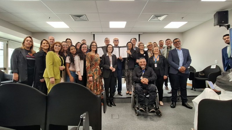 Fotografia de grupo feita em uma sala ampla e iluminada, semelhante a uma sala de reuni&otilde;es ou audit&oacute;rio. Cerca de duas dezenas de pessoas est&atilde;o reunidas no centro da imagem, posicionadas em duas fileiras e voltadas para a c&acirc;mera. Algumas pessoas, no centro do grupo, seguram folhas de papel brancas abertas, representando os termos assinados pelas pessoas presentes e voltadas para a fotografia.

Na parte central da frente, uma pessoa est&aacute; em uma cadeira de rodas, com as m&atilde;os unidas &agrave; frente do corpo, que &eacute; o presidente da FADERS, Marquinhos Lang. Ao redor, as demais pessoas est&atilde;o em p&eacute;. O grupo usa roupas variadas, incluindo ternos e blazers em tons escuros, al&eacute;m de pe&ccedil;as nas cores verde, marrom, branco e preto. Algumas pessoas utilizam crach&aacute;s.

Em primeiro plano, v&aacute;rias poltronas pretas vazias ocupam a parte inferior da fotografia e encobrem parcialmente a vis&atilde;o do grupo. &Agrave; direita, uma pessoa est&aacute; sentada em uma dessas poltronas, de costas e de perfil para a c&acirc;mera, segurando um objeto semelhante a um celular.

A sala possui carpete cinza, paredes claras e teto com lumin&aacute;rias retangulares e sa&iacute;das de ventila&ccedil;&atilde;o. Nas laterais superiores existem caixas de som pretas. &Agrave; esquerda h&aacute; grandes janelas, por onde entra luz natural. Ao fundo, atr&aacute;s do grupo, encontra-se uma grande tela de proje&ccedil;&atilde;o branca recolhida. &Agrave; direita, um monitor grande fixado &agrave; parede exibe uma interface de computador com textos pequenos, que n&atilde;o podem ser lidos na fotografia.

A composi&ccedil;&atilde;o concentra a aten&ccedil;&atilde;o no grupo reunido e, especialmente, nos documentos exibidos no centro.