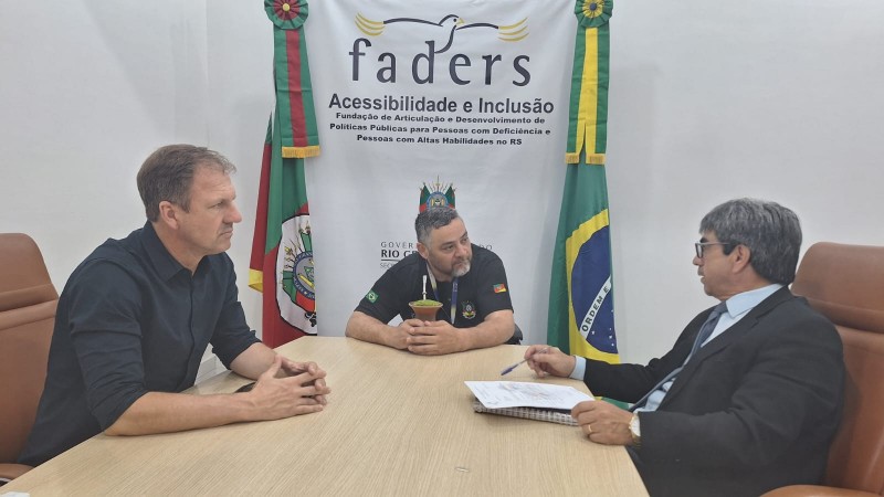 Fotografia registrada em uma sala de reuni&otilde;es da FADERS. Tr&ecirc;s pessoas est&atilde;o sentadas ao redor de uma mesa retangular de madeira clara. Ao centro da imagem, uma pessoa veste camiseta preta com identifica&ccedil;&atilde;o institucional e est&aacute; posicionada de frente para a c&acirc;mera, segurando uma cuia de chimarr&atilde;o sobre a mesa. &Agrave; esquerda, outra pessoa usa camisa escura de mangas compridas e mant&eacute;m as m&atilde;os apoiadas sobre a mesa, voltada para o centro da conversa. &Agrave; direita, uma terceira pessoa veste terno escuro, camisa clara e gravata, com um bloco de anota&ccedil;&otilde;es e uma caneta sobre a mesa.

Ao fundo, na parede branca, aparece o logotipo da FADERS e a inscri&ccedil;&atilde;o &ldquo;FADERS &ndash; Acessibilidade e Inclus&atilde;o&rdquo;. Abaixo, l&ecirc;-se: &ldquo;Funda&ccedil;&atilde;o de Articula&ccedil;&atilde;o e Desenvolvimento de Pol&iacute;ticas P&uacute;blicas para Pessoas com Defici&ecirc;ncia e Pessoas com Altas Habilidades no RS&rdquo;. Duas bandeiras est&atilde;o posicionadas atr&aacute;s da mesa: a bandeira do Rio Grande do Sul &agrave; esquerda e a bandeira do Brasil &agrave; direita.

Conforme informado, no centro est&aacute; Marquinhos Lang, presidente da FADERS, reunido com dois vereadores de Quara&iacute;, que ocupam os assentos &agrave; esquerda e &agrave; direita da mesa. O ambiente transmite o contexto de uma reuni&atilde;o institucional voltada ao di&aacute;logo e ao trabalho conjunto entre a funda&ccedil;&atilde;o e representantes do munic&iacute;pio