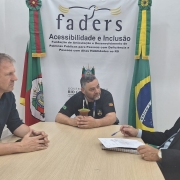 Fotografia registrada em uma sala de reuni&otilde;es da FADERS. Tr&ecirc;s pessoas est&atilde;o sentadas ao redor de uma mesa retangular de madeira clara. Ao centro da imagem, uma pessoa veste camiseta preta com identifica&ccedil;&atilde;o institucional e est&aacute; posicionada de frente para a c&acirc;mera, segurando uma cuia de chimarr&atilde;o sobre a mesa. &Agrave; esquerda, outra pessoa usa camisa escura de mangas compridas e mant&eacute;m as m&atilde;os apoiadas sobre a mesa, voltada para o centro da conversa. &Agrave; direita, uma terceira pessoa veste terno escuro, camisa clara e gravata, com um bloco de anota&ccedil;&otilde;es e uma caneta sobre a mesa.

Ao fundo, na parede branca, aparece o logotipo da FADERS e a inscri&ccedil;&atilde;o &ldquo;FADERS &ndash; Acessibilidade e Inclus&atilde;o&rdquo;. Abaixo, l&ecirc;-se: &ldquo;Funda&ccedil;&atilde;o de Articula&ccedil;&atilde;o e Desenvolvimento de Pol&iacute;ticas P&uacute;blicas para Pessoas com Defici&ecirc;ncia e Pessoas com Altas Habilidades no RS&rdquo;. Duas bandeiras est&atilde;o posicionadas atr&aacute;s da mesa: a bandeira do Rio Grande do Sul &agrave; esquerda e a bandeira do Brasil &agrave; direita.

Conforme informado, no centro est&aacute; Marquinhos Lang, presidente da FADERS, reunido com dois vereadores de Quara&iacute;, que ocupam os assentos &agrave; esquerda e &agrave; direita da mesa. O ambiente transmite o contexto de uma reuni&atilde;o institucional voltada ao di&aacute;logo e ao trabalho conjunto entre a funda&ccedil;&atilde;o e representantes do munic&iacute;pio